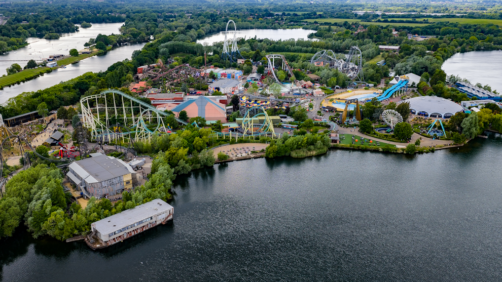 A high-resolution drone photograph of Thorpe Park amusement park in Thorpe Village, Surrey, showing rollercoasters, rides, and surrounding lakes